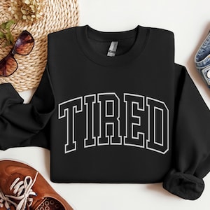 Pu&ograve; includere: Felpa nera girocollo con la scritta "TIRED" a grandi lettere bianche. La felpa &egrave; esposta con occhiali da sole, scarpe marroni e un paio di jeans.