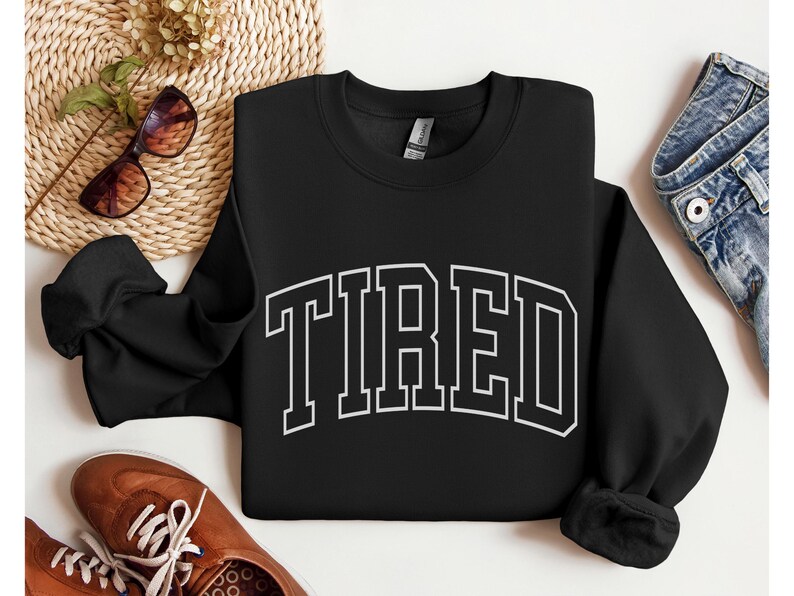 Pu&ograve; includere: Felpa nera girocollo con la scritta "TIRED" a grandi lettere bianche. La felpa &egrave; esposta con occhiali da sole, scarpe marroni e un paio di jeans.