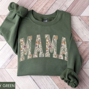 May include: Military green sweatshirt with the word "MAMA" in a floral design. The letters are filled with a pattern of flowers in shades of pink, orange, and cream. The sweatshirt has long sleeves and a crew neck.