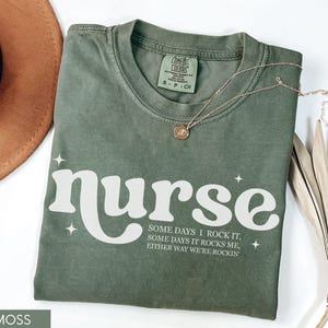 Pode incluir: Camiseta verde musgo com a palavra "nurse" em branco, juntamente com o texto "Some days I rock it, some days it rocks me, either way we're rockin'". Um colar dourado e um chap&eacute;u castanho tamb&eacute;m s&atilde;o vis&iacute;veis.