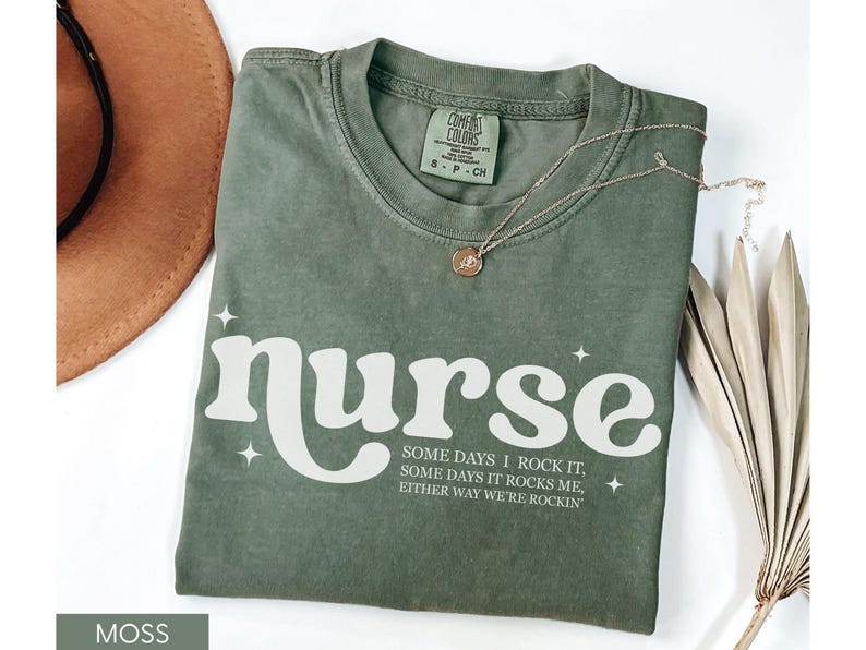 Pode incluir: Camiseta verde musgo com a palavra "nurse" em branco, juntamente com o texto "Some days I rock it, some days it rocks me, either way we're rockin'". Um colar dourado e um chap&eacute;u castanho tamb&eacute;m s&atilde;o vis&iacute;veis.