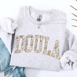 May include: A light gray sweatshirt with the word "DOULA" in large, floral-patterned letters across the front. The sweatshirt is folded, with the sleeves visible, and is paired with blue jeans. A silver necklace with a small pendant rests on the sweatshirt.