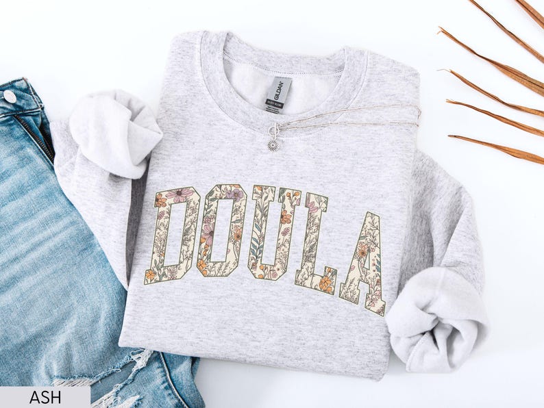 May include: A light gray sweatshirt with the word "DOULA" in large, floral-patterned letters across the front. The sweatshirt is folded, with the sleeves visible, and is paired with blue jeans. A silver necklace with a small pendant rests on the sweatshirt.
