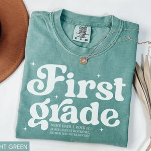 Puede incluir: Camiseta verde claro con las palabras "First Grade" en letras blancas grandes. La camiseta tambi&eacute;n tiene el texto "SOME DAYS I ROCK IT, SOME DAYS IT ROCKS ME, EITHER WAY WE'RE ROCKIN" en letras blancas m&aacute;s peque&ntilde;as. Un collar de oro est&aacute; en la camiseta.