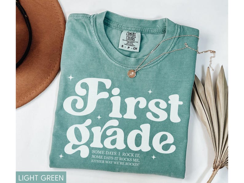Puede incluir: Camiseta verde claro con las palabras "First Grade" en letras blancas grandes. La camiseta tambi&eacute;n tiene el texto "SOME DAYS I ROCK IT, SOME DAYS IT ROCKS ME, EITHER WAY WE'RE ROCKIN" en letras blancas m&aacute;s peque&ntilde;as. Un collar de oro est&aacute; en la camiseta.