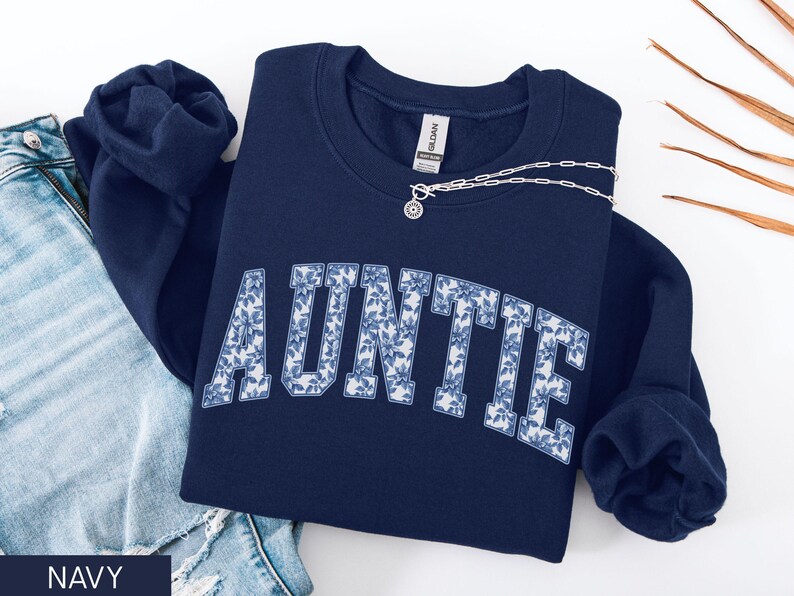 Peut inclure: Sweat-shirt bleu marine avec le mot "AUNTIE" en lettres blanches remplies d'un motif floral bleu. Un collier argent&eacute; avec un pendentif est sur le sweat-shirt. Le mot "NAVY" est en bas.