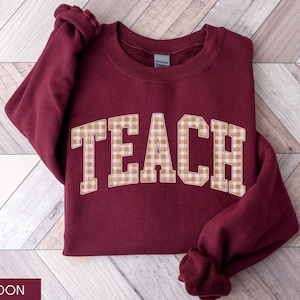 May include: Maroon sweatshirt with the word "TEACH" in large, tan and white checkered letters across the front. The sweatshirt is folded on a light brown and white wooden surface. The sleeves are folded.