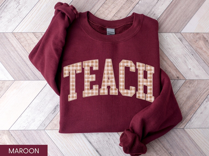 May include: Maroon sweatshirt with the word "TEACH" in large, tan and white checkered letters across the front. The sweatshirt is folded on a light brown and white wooden surface. The sleeves are folded.