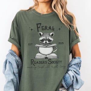 May include: A green t-shirt with a raccoon reading a book and the text "Feral Readers Society est. 2025 reading trash all night long".