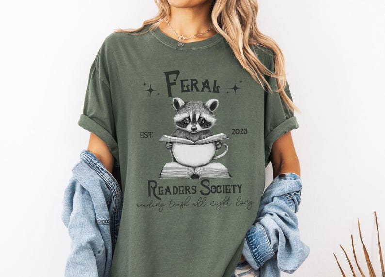 May include: A green t-shirt with a raccoon reading a book and the text "Feral Readers Society est. 2025 reading trash all night long".