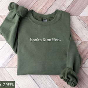 May include: Military green sweatshirt with the text "books & coffee" in white. The long-sleeved crewneck top is made of a soft material and is displayed on a wooden surface. The sleeves are gathered at the wrists.