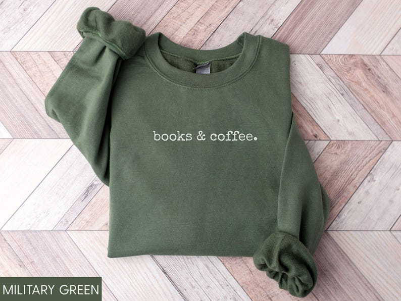 May include: Military green sweatshirt with the text "books & coffee" in white. The long-sleeved crewneck top is made of a soft material and is displayed on a wooden surface. The sleeves are gathered at the wrists.