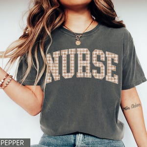 May include: A dark gray t-shirt with the word "NURSE" in large, block letters across the chest. The letters have a tan and white plaid pattern. The shirt is a casual style.