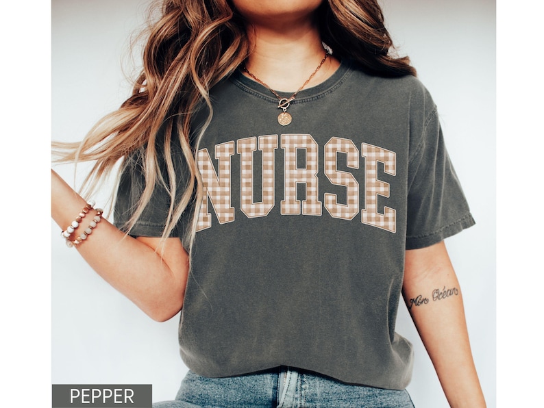 May include: A dark gray t-shirt with the word "NURSE" in large, block letters across the chest. The letters have a tan and white plaid pattern. The shirt is a casual style.