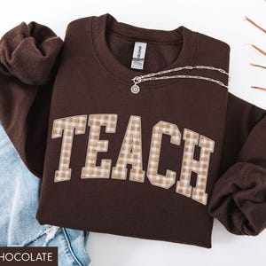 May include: A dark chocolate-colored sweatshirt with the word "TEACH" in a plaid pattern across the front. A silver necklace with a pendant is draped across the sweatshirt. The sweatshirt is paired with blue jeans.
