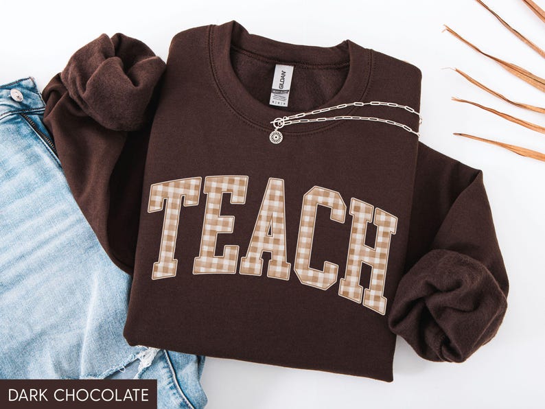 May include: A dark chocolate-colored sweatshirt with the word "TEACH" in a plaid pattern across the front. A silver necklace with a pendant is draped across the sweatshirt. The sweatshirt is paired with blue jeans.