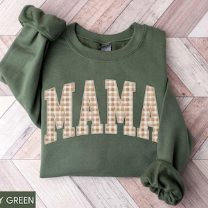 May include: A military green sweatshirt with the word "MAMA" in large, tan and white gingham letters across the front. The sweatshirt has long sleeves with ribbed cuffs. The bottom of the sweatshirt is also ribbed.