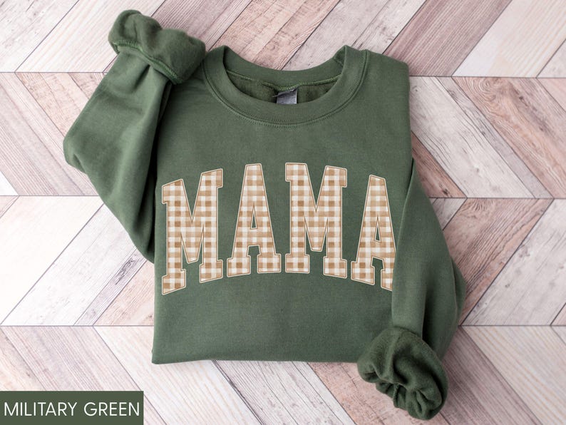 May include: A military green sweatshirt with the word "MAMA" in large, tan and white gingham letters across the front. The sweatshirt has long sleeves with ribbed cuffs. The bottom of the sweatshirt is also ribbed.