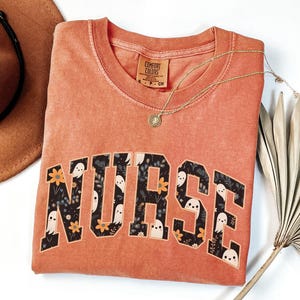 May include: A burnt orange t-shirt with the word "NURSE" printed in black letters. The letters are filled with a pattern of white ghosts and orange flowers on a black background.