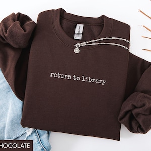 May include: Dark chocolate-colored sweatshirt with the phrase "return to library" embroidered in white. A silver necklace with a circular pendant rests on the sweatshirt. Light blue denim jeans are visible in the background.