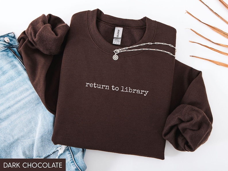 May include: Dark chocolate-colored sweatshirt with the phrase "return to library" embroidered in white. A silver necklace with a circular pendant rests on the sweatshirt. Light blue denim jeans are visible in the background.
