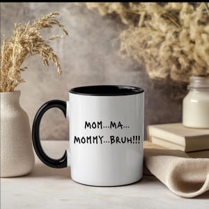 May include: White ceramic mug with a black interior and handle. The mug features the text "MOM...MA... MOMMY...BRUH!!!" in black lettering. The mug is displayed on a light-colored surface with decorative elements in the background.