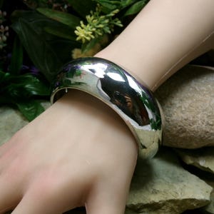 May include: A wide, polished silver bangle bracelet. The bracelet is smooth and reflective, with a rounded shape. The light reflects off the surface, creating a bright, shiny appearance. The bracelet is worn on a wrist.