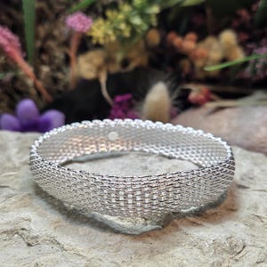 May include: A silver-toned bangle bracelet featuring a fine, flexible mesh construction with a woven, chain-link texture. The bracelet has a rounded, continuous band design with a polished metallic finish that reflects light across its surface.