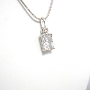 May include: A silver pendant necklace featuring a rectangular, clear gemstone. The pendant is suspended from a delicate silver chain. The gemstone is set in a silver frame, and a smaller, round stone sits above it.
