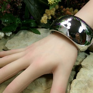 May include: A wide, polished silver cuff bracelet. The bracelet reflects the surrounding environment, including greenery and a camera. The bracelet is worn on a pale arm, with a background of rocks and flowers. The cuff is a statement piece of jewelry.