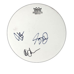 May include: White drumhead with three autographs in blue ink. The drumhead is a Remo Weather King Ambassador Batter, made in the USA.