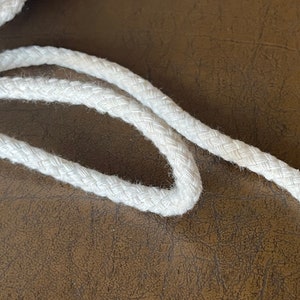May include: White cotton rope with a braided texture. The rope is coiled on a brown surface.