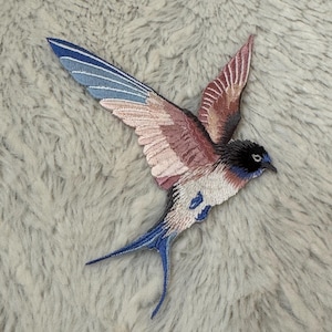 May include: Embroidered patch of a swallow bird with outstretched wings. The bird features a black head, white chest, and a blue and pink wing design. The tail is blue, and the patch is on a gray, fuzzy background.