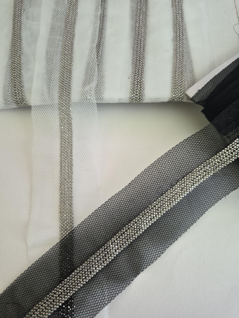 May include: Close-up of decorative trim featuring silver beads on white and black mesh fabric. The trim is arranged in vertical stripes on the white fabric and a single strip on the black fabric. The mesh has a hexagonal pattern.