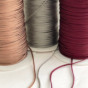 May include: Three spools of elastic cord in different colors: rose gold, gray, and burgundy. The cords are wound around the spools and are ready to be used for crafting projects.