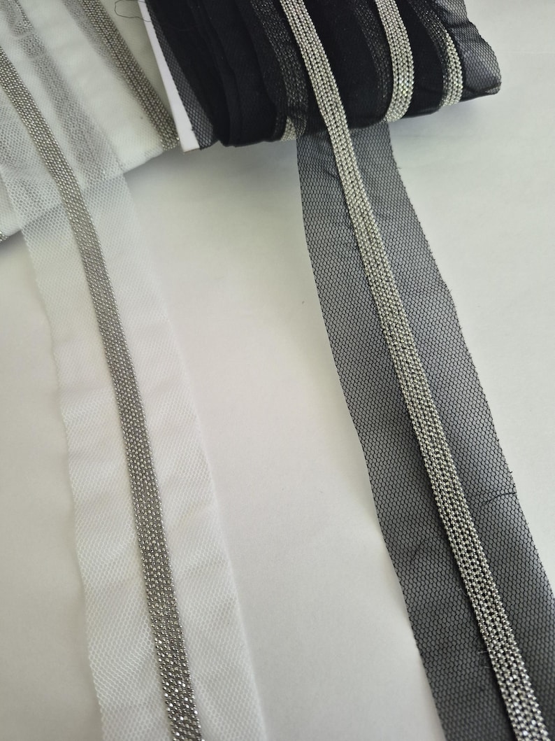 May include: Rolls of black and white tulle ribbon with silver beaded trim. The ribbon is displayed on a white surface. The tulle has a mesh-like appearance, and the silver trim adds a decorative element to the ribbon.