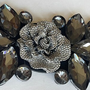 May include: A silver-toned rose embellishment surrounded by black rhinestones. The rose is detailed with a textured surface and is a popular embellishment for clothing, accessories, and crafts.
