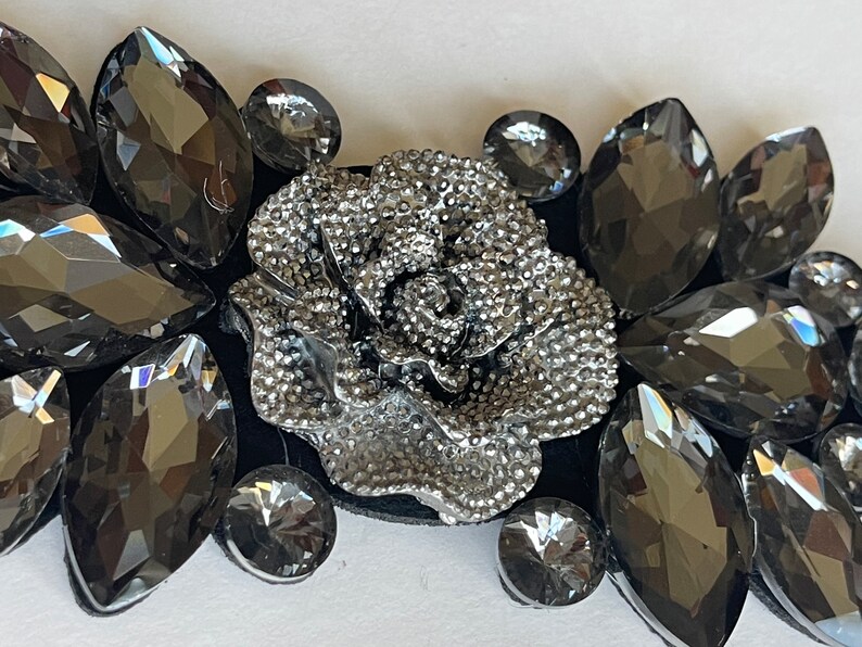 May include: A silver-toned rose embellishment surrounded by black rhinestones. The rose is detailed with a textured surface and is a popular embellishment for clothing, accessories, and crafts.