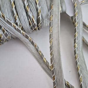 May include: A spool of silver-gray decorative trim with a gold and white twisted rope design. The trim is made of a soft, woven material and is ideal for embellishing clothing, accessories, or home decor projects.