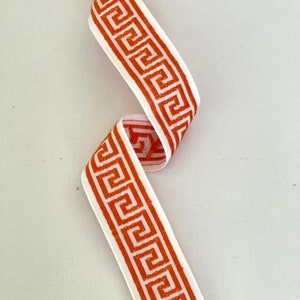 May include: Orange and white ribbon with a geometric pattern. The pattern is a repeating Greek key design.