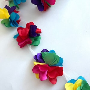 May include: A colorful garland of fabric flowers, each flower is made of multiple layers of fabric in different colors, including red, yellow, green, blue, and purple. The flowers are attached to a white mesh ribbon.