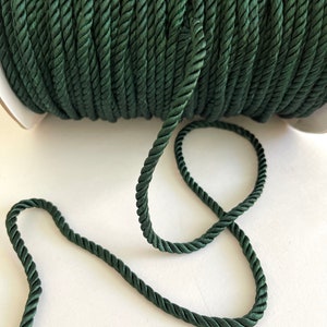 May include: A spool of dark green twisted rope. The rope is tightly wound on a white cardboard spool. A length of the rope extends from the spool, showing its texture and color. The rope is a deep, forest green.
