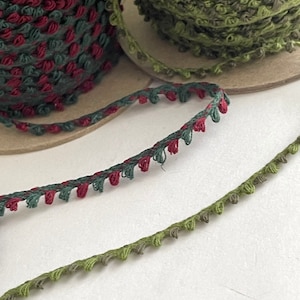 May include: Close-up of spools of decorative trim in shades of green and burgundy. The trim has a looped edge, and is wound on cardboard spools. The trim is suitable for sewing, crafting, and embellishing various projects.