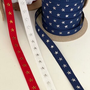 May include: Spools of ribbon in red, white, and navy blue, each adorned with star patterns. The red ribbon features gold stars, the white ribbon has silver stars, and the navy ribbon has silver stars. Ideal for crafting and decorative projects.