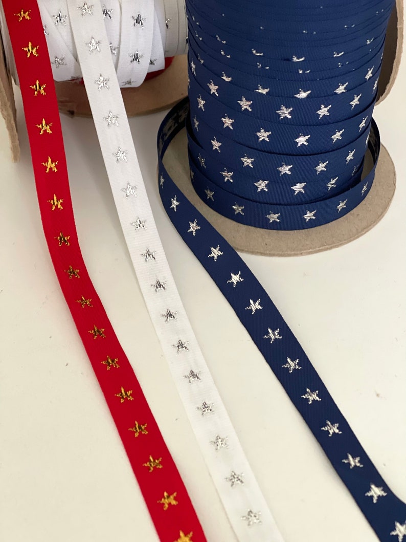 May include: Spools of ribbon in red, white, and navy blue, each adorned with star patterns. The red ribbon features gold stars, the white ribbon has silver stars, and the navy ribbon has silver stars. Ideal for crafting and decorative projects.