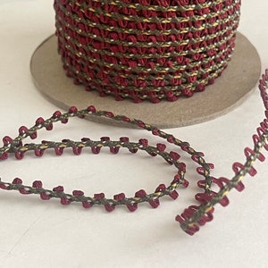 May include: A spool of burgundy and olive green trim with a decorative stitch. The trim is made of a woven fabric and has a slightly textured surface.