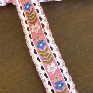 May include: Pink and white crocheted trim with a floral design featuring blue, pink, and yellow flowers. The trim has a delicate, lacy pattern.