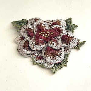 May include: A close-up of a burgundy and white embroidered flower patch with green leaves. The flower has a layered design and is detailed with gold stitching.