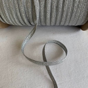 May include: A spool of silver elastic trim with a metallic sheen. The trim is approximately 1/4 inch wide and has a subtle ribbed texture.