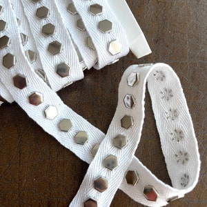 May include: White ribbon with silver hexagonal studs. The ribbon is tightly woven and has a zig-zag pattern. The studs are evenly spaced along the ribbon's length, creating a decorative trim. The ribbon is on a brown surface.